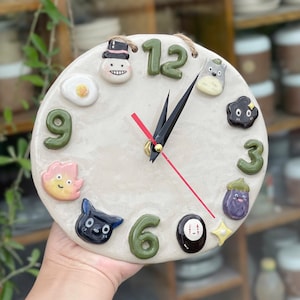 May include: A handmade ceramic clock with a cream-coloured face. The clock features whimsical, colourful, raised designs in place of numbers, including a fried egg, a smiling face, and a black cat. The clock hands are black, with a red second hand.