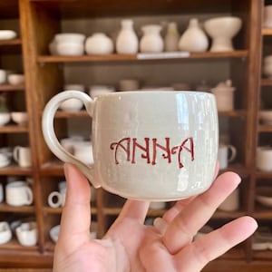 Puede incluir: Una taza de cer&aacute;mica beige con un asa curvada. El nombre "ANNA" est&aacute; escrito en letras rojas y estilizadas en la parte delantera de la taza. La taza se sostiene en una mano, con una estanter&iacute;a de madera llena de cer&aacute;mica al fondo.
