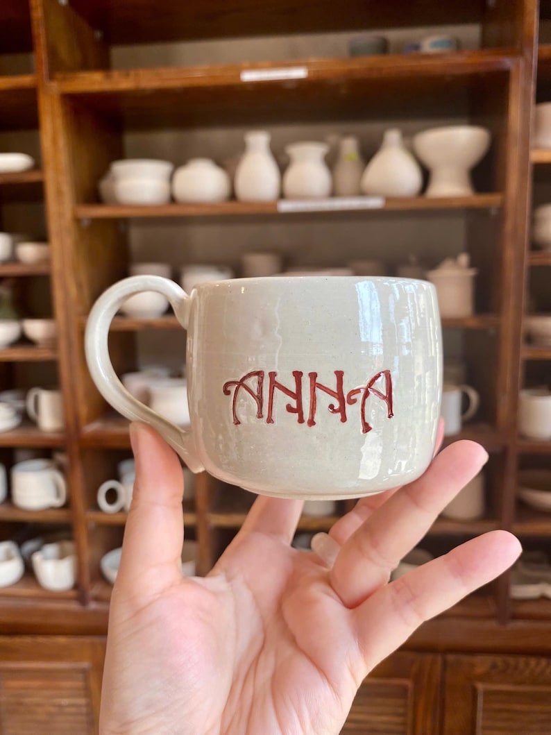 Puede incluir: Una taza de cer&aacute;mica beige con un asa curvada. El nombre "ANNA" est&aacute; escrito en letras rojas y estilizadas en la parte delantera de la taza. La taza se sostiene en una mano, con una estanter&iacute;a de madera llena de cer&aacute;mica al fondo.