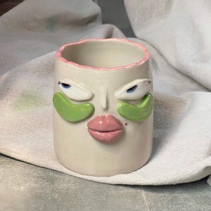May include: A ceramic mug featuring a sculpted face with a cream-colored base, a wavy pink rim, and prominent pink lips. The face includes two white eyes with blue pupils and green under-eye patches applied to the cheeks, with a small red dot.