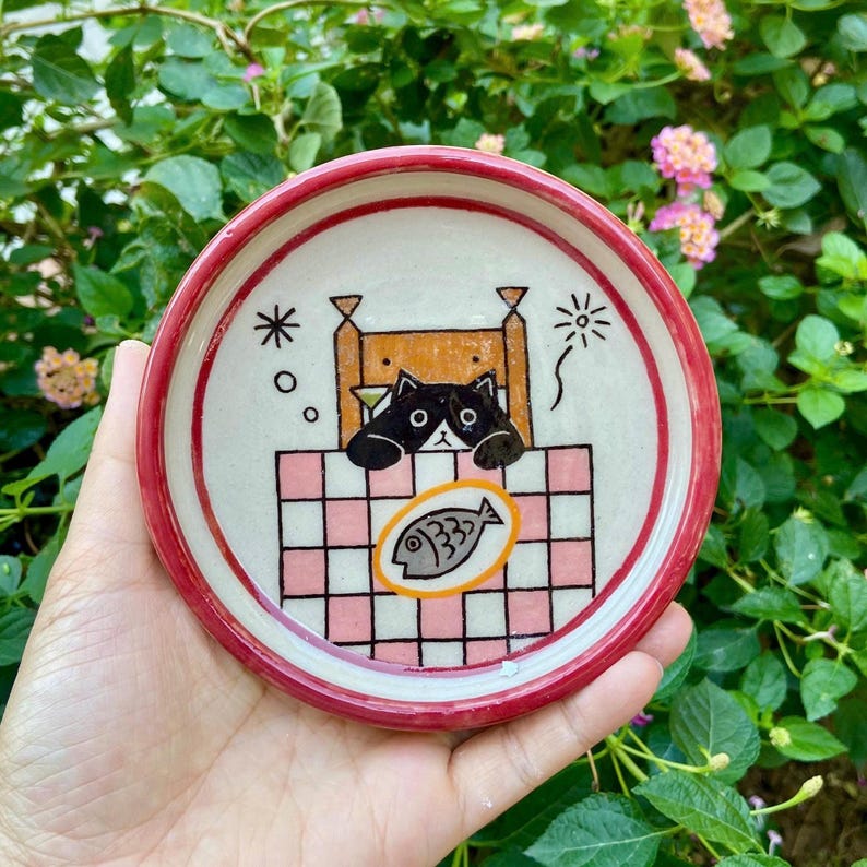 May include: A round ceramic dish with a red rim featuring a hand-painted illustration of a black and white cat lying in a bed with a pink and white checkered blanket. A gray fish icon is centered on the blanket, with star and firework accents.