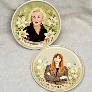 Puede incluir: Dos platos redondos de cerámica con retratos pintados a mano. El plato superior muestra a una persona rubia con top negro rodeada de lirios blancos con el texto "Толстухина Т.В.". El inferior muestra a una pelirroja con blazer de cuadros, margaritas y el texto "Аристархова Т.А.".