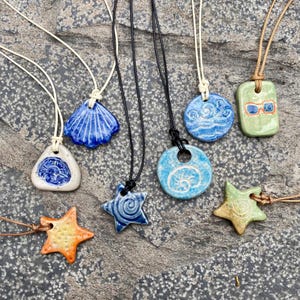 May include: Seven ceramic pendants on cords, each with a different design. The pendants include a seashell, a starfish, a circle with a wave design, a circle with a spiral design, a circle with a seashell design, a starfish with a spiral design, and a rectangle with a pair of sunglasses design.