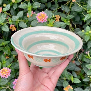 May include: A handmade ceramic bowl with a cream interior and a light blue spiral design. The rim features orange fish illustrations. The bowl is held against a backdrop of green foliage and pink flowers.