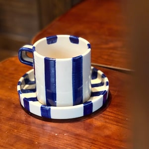 Könnte beinhalten: Eine Kaffeetasse aus Keramik mit passender Untertasse, verziert mit einem handgemalten Muster aus kräftigen, vertikalen marineblauen Streifen auf cremeweißem Grund. Die Tasse hat einen stabilen, eckigen Henkel und die Untertasse einen gestreiften Rand.