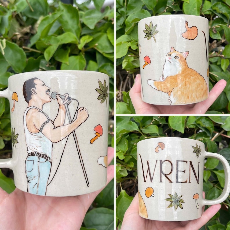 May include: Three ceramic mugs with unique designs. One mug features a singer with a microphone, another has an orange cat, and the third displays the name "WREN". All mugs have mushroom and leaf illustrations.