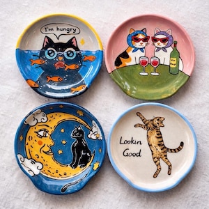 May include: Four ceramic spoon rests with cat illustrations. One shows a black cat in water with the text "I'm hungry". Another depicts two cats with sunglasses and wine. A third shows a cat on a moon, and the last says "Lookin Good".