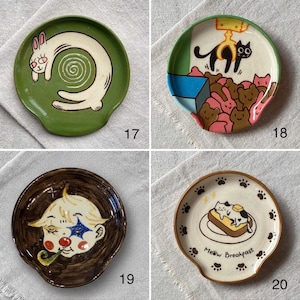 K&ouml;nnte beinhalten: Vier Keramikplatten mit einzigartigen Designs. Eine zeigt ein wei&szlig;es Kaninchen auf gr&uuml;nem Grund, eine andere schwarze Katzen und rosa B&auml;ren, eine dritte einen Clown und die letzte sagt "Meow Breakfast" mit einer Katze auf Toast.
