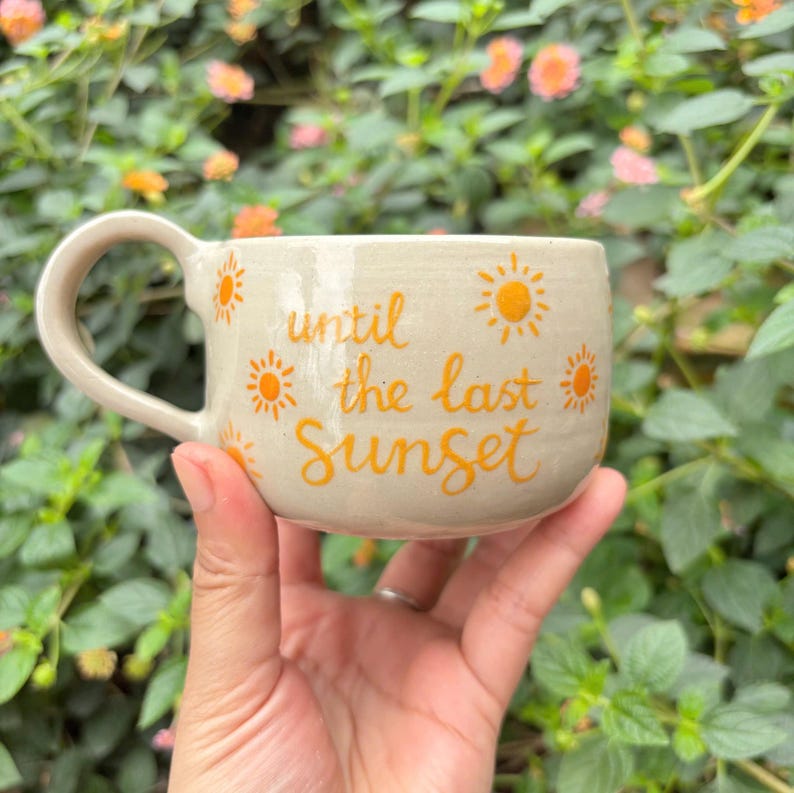 Puede incluir: Una taza de cer&aacute;mica beige con un asa curva. La taza est&aacute; decorada con ilustraciones de sol naranja y las palabras "until the last Sunset" en escritura naranja. La taza se sostiene en una mano.