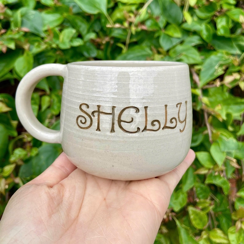 Puede incluir: Una taza de cer&aacute;mica color crema con el nombre "SHELLY" en letras marrones. La taza tiene una forma redondeada con un asa grande y l&iacute;neas horizontales texturizadas. La taza se sostiene en una mano sobre un fondo de follaje verde.