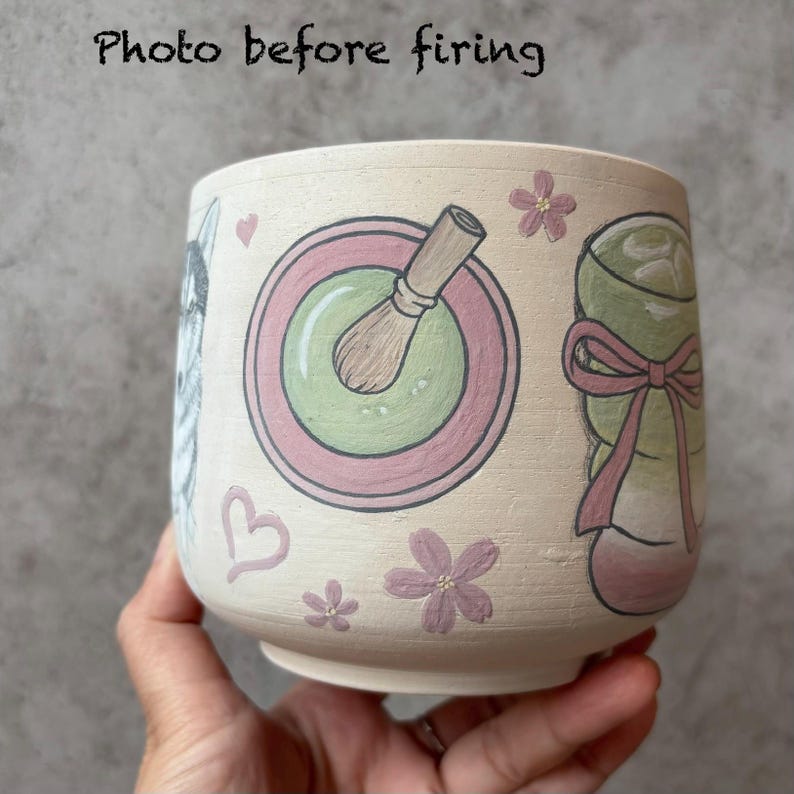 May include: A beige ceramic cup with hand-painted illustrations. The cup features a matcha bowl with a whisk, a wrapped gift, a dog, hearts, and flowers. The text "Photo before firing" is at the top.