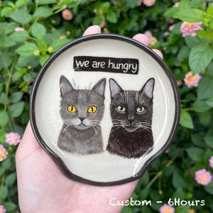 K&ouml;nnte beinhalten: Ein Keramik-L&ouml;ffelhalter mit zwei Katzenportr&auml;ts. Eine Katze ist grau mit gelben Augen, die andere ist schwarz mit gr&uuml;nen Augen. Der L&ouml;ffelhalter hat einen schwarzen Rand und den Text "we are hungry" oben.