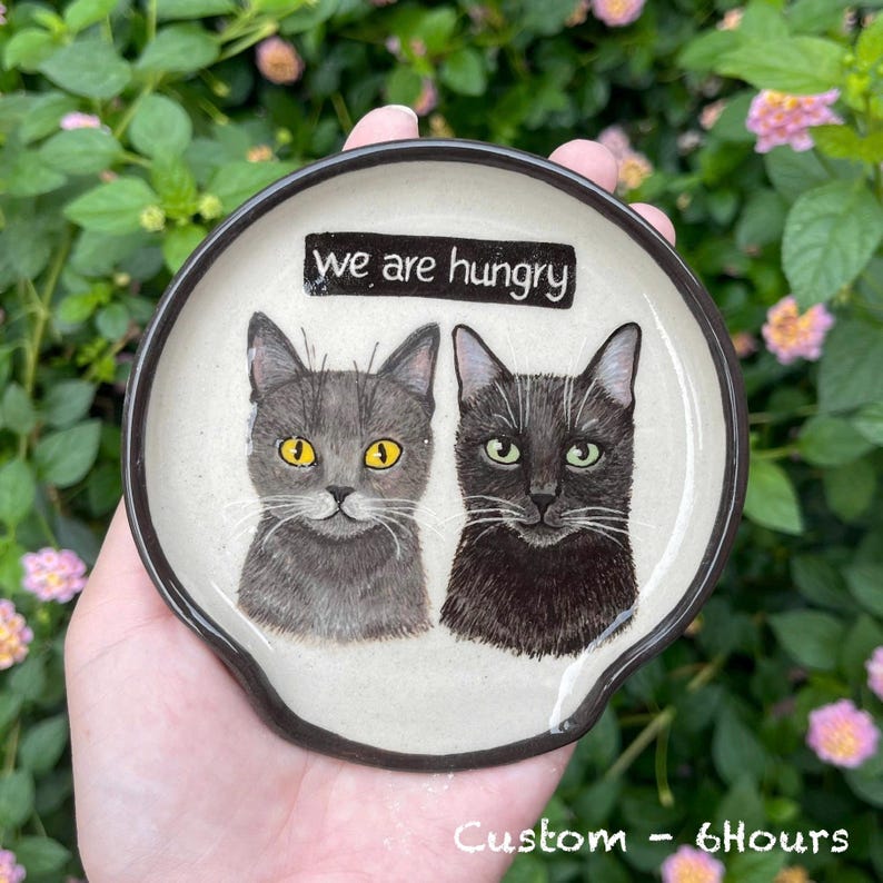 K&ouml;nnte beinhalten: Ein Keramik-L&ouml;ffelhalter mit zwei Katzenportr&auml;ts. Eine Katze ist grau mit gelben Augen, die andere ist schwarz mit gr&uuml;nen Augen. Der L&ouml;ffelhalter hat einen schwarzen Rand und den Text "we are hungry" oben.