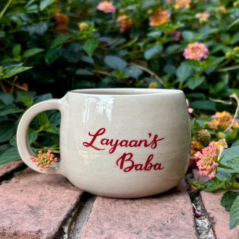 Puede incluir: Una taza de cer&aacute;mica beige con forma redondeada y un asa grande. La taza presenta el texto rojo "Layaan's Baba" en su lateral. El fondo incluye follaje verde y flores rosas y amarillas.
