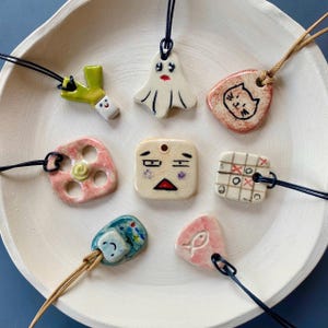 Könnte beinhalten: Eine Sammlung handgefertigter Keramik-Anhänger-Halsketten mit verschiedenen Designs, darunter ein Geist, eine Katze und ein Tic-Tac-Toe-Spiel. Jeder Anhänger ist an einer schwarzen oder beigefarbenen Kordel befestigt. Die Anhänger werden auf einer weißen, flachen Schale präsentiert.