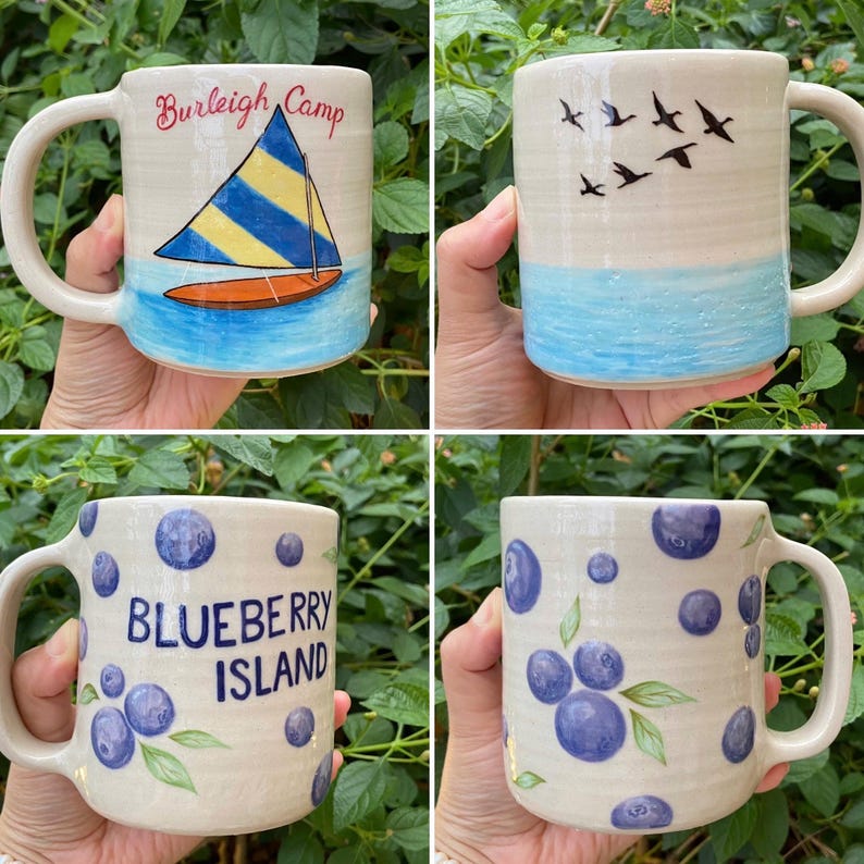 May include: Four ceramic mugs with unique designs. One mug features a sailboat with the text "Burleigh Camp." Another depicts birds flying over blue water. The other two mugs have a blueberry theme, one with the text "Blueberry Island."