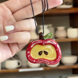 Può includere: Ciondolo a forma di mela in ceramica con cordino nero. La mela è rossa con dettagli a stella bianchi e un centro giallo, con due dettagli di semi marroni. Una foglia verde e un gambo marrone completano il design. Il ciondolo è tenuto contro uno sfondo sfocato di ceramica.