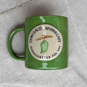 Könnte beinhalten: Ein grüner Keramikbecher mit einem kreisförmigen, cremefarbenen Emblem auf der Vorderseite. Das Emblem zeigt eine gemalte grüne Puppe auf einem braunen Zweig mit dem schwarzen Text CHRYSALIS APOTHECARY HANDCRAFTED FOR YOU am Rand.