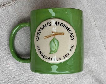 Könnte beinhalten: Ein grüner Keramikbecher mit einem cremefarbenen, kreisförmigen Emblem auf der Vorderseite. Das Emblem zeigt einen braunen Zweig mit einer grünen Puppe und den schwarzen Schriftzug CHRYSALIS APOTHECARY HANDCRAFTED FOR YOU im Kreis.