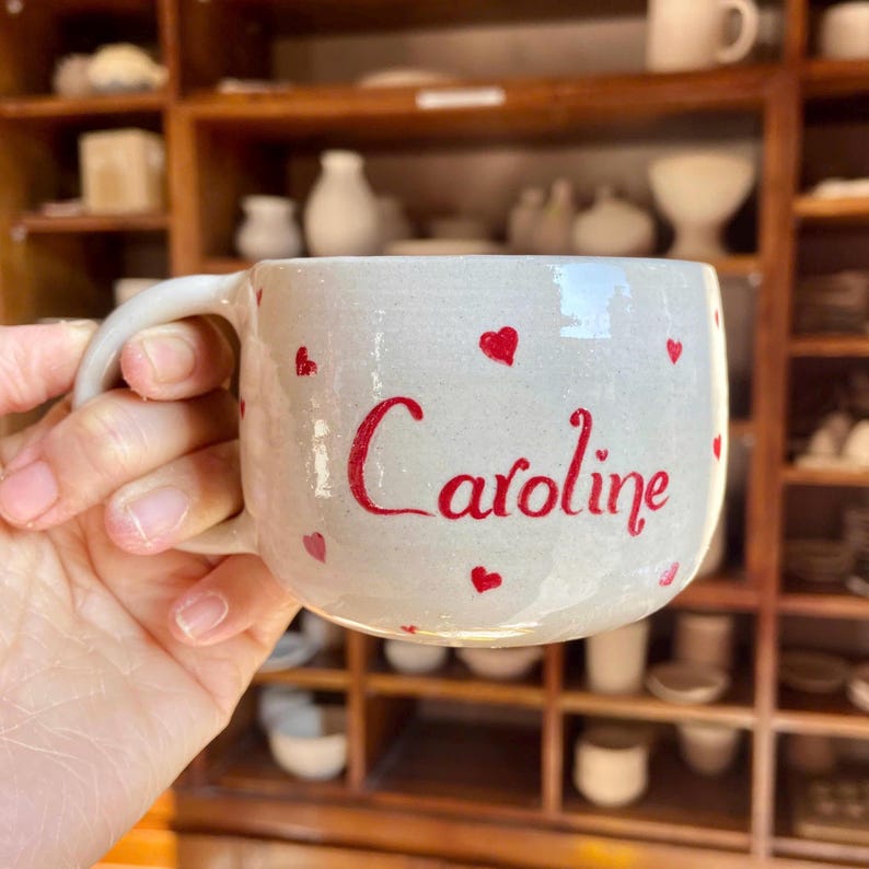 Puede incluir: Taza de cer&aacute;mica hecha a mano con base blanca, adornada con corazones rojos y el nombre "Caroline" en escritura roja. La taza tiene un asa curva. Fondo: estanter&iacute;as de madera llenas de cer&aacute;mica.