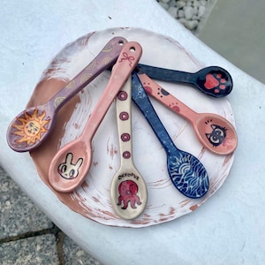 Puede incluir: Cinco cucharas de cerámica dispuestas sobre un plato redondo de color blanco y terracota. Las cucharas presentan diseños pintados a mano que incluyen un sol, un conejo, un pulpo con el texto OCTOPUS, ondas azules y una cara de gato.