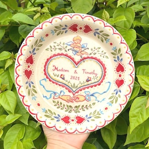 May include: An off-white decorative plate with a heart-shaped design. It features the names "Madison & Timothy" and the year "2021." The plate is decorated with red hearts, blue ribbons, and a cherub with a bow and arrow. The rim has a scalloped edge.