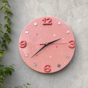 Puede incluir: Un reloj de pared redondo de color rosa con números en relieve en las posiciones de las 12, 3, 6 y 9. La esfera está decorada con pequeñas estrellas y puntos blancos y rosas, con marcadores circulares plateados y manecillas negras.