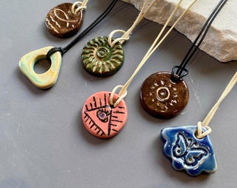 May include: A collection of six ceramic pendants on cords. The pendants feature various shapes and colours, including a brown fish design, a green sun, a pink eye, a brown sun, a blue butterfly, and a light green triangle with a central hole.