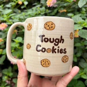 May include: A cream-coloured ceramic mug featuring the phrase "Tough Cookies" in brown. The mug is adorned with hand-painted chocolate chip cookie designs. The mug has a large handle and is held in a person's hand.