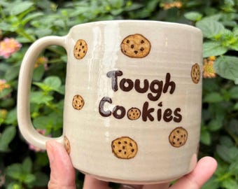 May include: A beige ceramic mug with the words "Tough Cookies" in brown lettering. The mug is decorated with hand-painted chocolate chip cookies. The mug has a large handle and is held in a person's hand.