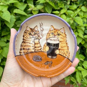 May include: A round ceramic spoon rest with a purple rim, featuring a hand-painted illustration of two orange tabby cats at a table. One cat holds a pint glass labeled Guinness, while the other sits near an ashtray and a small fish icon.