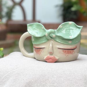 May include: A ceramic mug shaped like a face with closed eyes, pink cheeks, and pink lips. The mug features a light green headband with white polka dots tied in a knot at the top. It has a curved handle on the left side and a smooth finish.