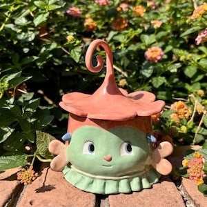 Może przedstawiać: Kapryśny ceramiczny słoik z pokrywką, z zieloną twarzą z niebieskimi oczami i różowym, kwiatowym wierzchem. Słoik ma falbankowy kołnierz, na tle zielonych liści i kwiatów.