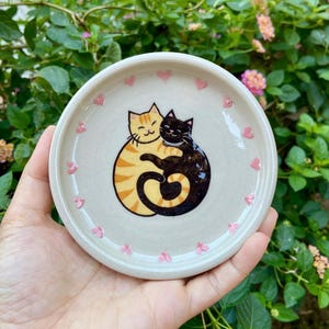 May include: A round, off-white ceramic trinket dish featuring a hand-painted illustration of an orange tabby cat and a black cat hugging each other. The cats' tails form a heart shape. Small, light pink painted hearts are scattered around the rim.