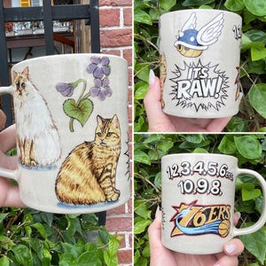 May include: Three ceramic mugs with various designs. One mug features a white cat and a tabby cat with purple flowers. Another mug has a cartoon turtle with wings and the text "ITS RAW!". The third mug displays numbers and the "76ERS" logo.
