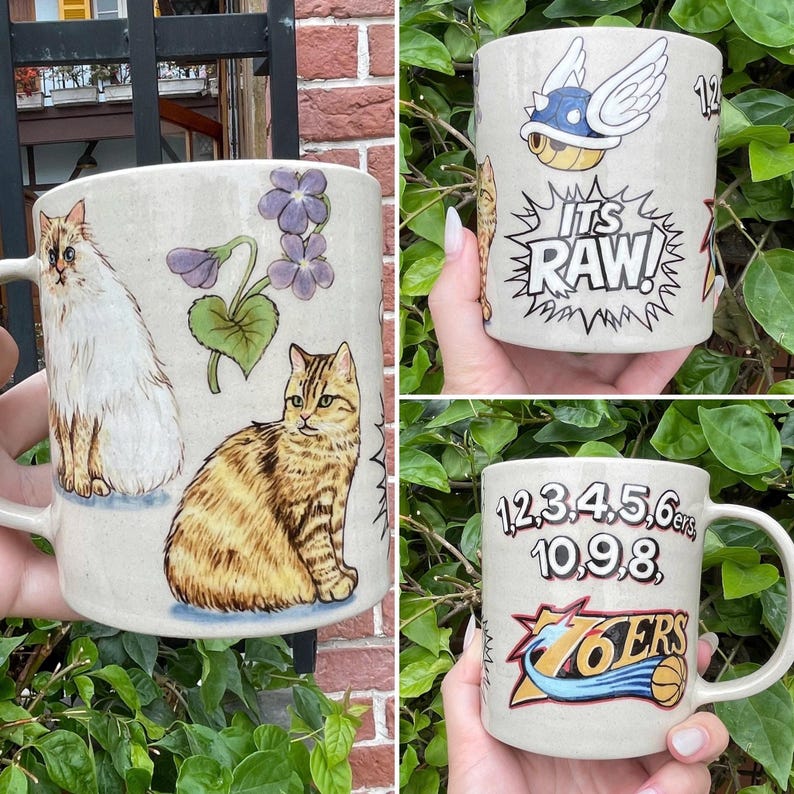 May include: Three ceramic mugs with various designs. One mug features a white cat and a tabby cat with purple flowers. Another mug has a cartoon turtle with wings and the text "ITS RAW!". The third mug displays numbers and the "76ERS" logo.