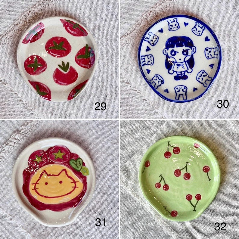 K&ouml;nnte beinhalten: Vier kleine Keramik-L&ouml;ffelablagen mit einzigartigen Designs. Eine zeigt rote Tomaten auf wei&szlig;em Grund, eine andere ein blau umrandetes M&auml;dchen-Design, eine dritte ein Katzen-Design und die letzte rote Kirschen auf gr&uuml;nem Grund. Jede hat einen kleinen Griff.