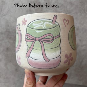May include: A hand-painted ceramic cup with a matcha drink design. The cup features a green matcha drink with ice, a pink bow, and floral accents. The text "Photo before firing" is at the top.