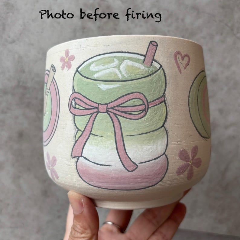 May include: A hand-painted ceramic cup with a matcha drink design. The cup features a green matcha drink with ice, a pink bow, and floral accents. The text "Photo before firing" is at the top.