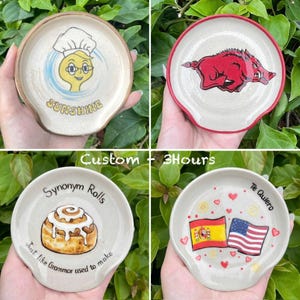 K&ouml;nnte beinhalten: Vier Keramik-L&ouml;ffelablagen mit einzigartigen Designs. Eine zeigt einen Koch mit dem Wort "Sunshine", eine andere ein rotes Wildschwein, eine dritte mit einer Zimtschnecke und dem Text "Synonym Rolls" und die letzte mit Flaggen und den Worten "Te Quiero".