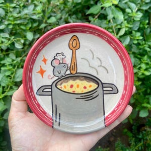 May include: A round ceramic dish with a red rim featuring a hand-painted illustration of a grey mouse wearing a chef hat, standing next to a large wooden spoon in a grey pot filled with yellow soup containing red and green vegetable pieces.