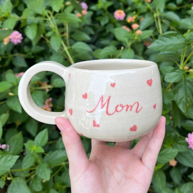 Puede incluir: Taza de cer&aacute;mica color crema con la palabra "Mom" en escritura roja, adornada con peque&ntilde;os corazones rojos. La taza tiene un asa redondeada y se sostiene sobre un fondo de follaje verde.