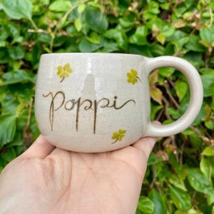 Puede incluir: Taza de cer&aacute;mica color crema con la palabra "poppi" en escritura marr&oacute;n. La taza presenta dise&ntilde;os de tr&eacute;boles amarillos y un asa grande. La taza se sostiene en una mano sobre un fondo de follaje verde.