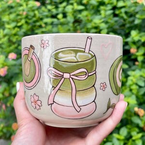 May include: A ceramic mug with a hand-painted matcha latte design. The mug features a light beige background with illustrations of a matcha drink, a whisk, flowers, and a pink bow. The design is in shades of green, pink, and white.