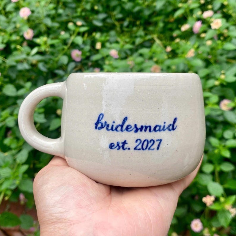 Puede incluir: Una taza de cer&aacute;mica beige con forma redondeada y asa curva. La taza tiene el texto azul "Bridesmaid est. 2027" en un lado. El fondo es una vista borrosa de follaje verde y flores rosas.