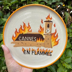 May include: A round ceramic plate with an orange rim featuring a hand-painted illustration of a building with a clock tower engulfed in orange and yellow flames. The text CANNES is printed on the building, and EN FLAMMES is below it.