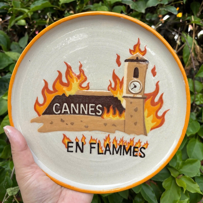 May include: A round ceramic plate with an orange rim featuring a hand-painted illustration of a building with a clock tower engulfed in orange and yellow flames. The text CANNES is printed on the building, and EN FLAMMES is below it.