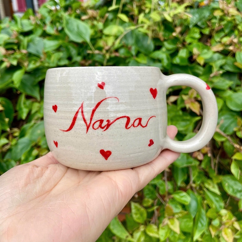 Puede incluir: Taza de cer&aacute;mica hecha a mano con base color crema, adornada con corazones rojos y el nombre "Narra" en escritura roja. La taza tiene forma redondeada y un asa grande.