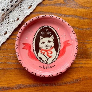 May include: A round pink ceramic trinket dish featuring a central dark brown oval frame containing a white cat illustration with a red bow. The dish has a decorative black border pattern, red ribbon accents, and the name bella written in black text.