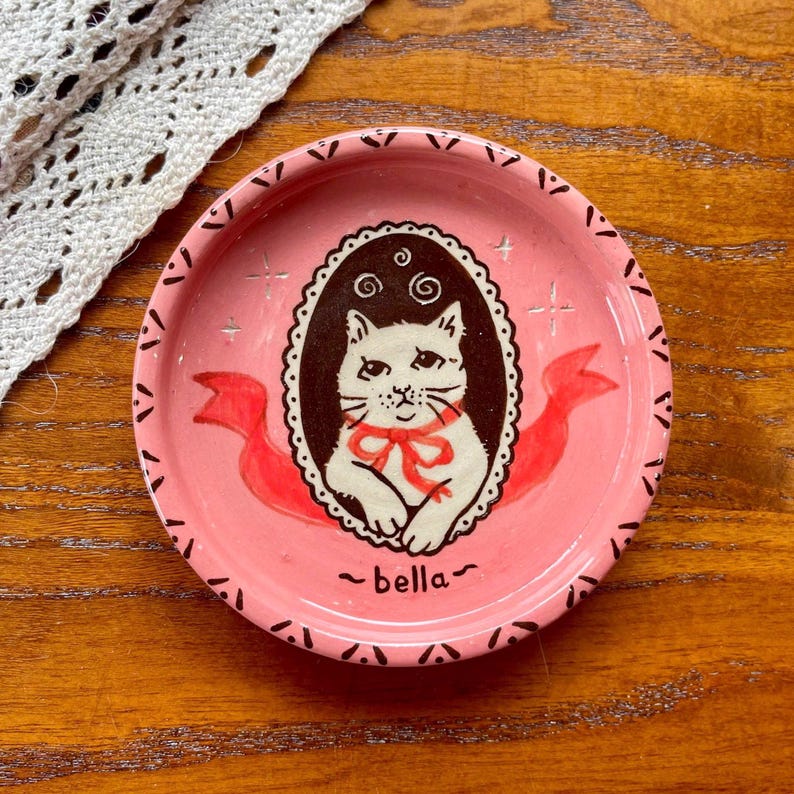 May include: A round pink ceramic trinket dish featuring a central dark brown oval frame containing a white cat illustration with a red bow. The dish has a decorative black border pattern, red ribbon accents, and the name bella written in black text.